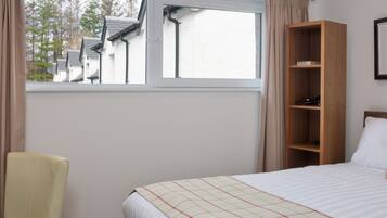 A bedroom with a bed, a wooden shelf, a chair, and a window with a view of houses and trees.