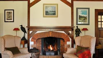 A cozy living room with a brick fireplace, two patterned armchairs, a table with a book, and framed artwork on the walls.
