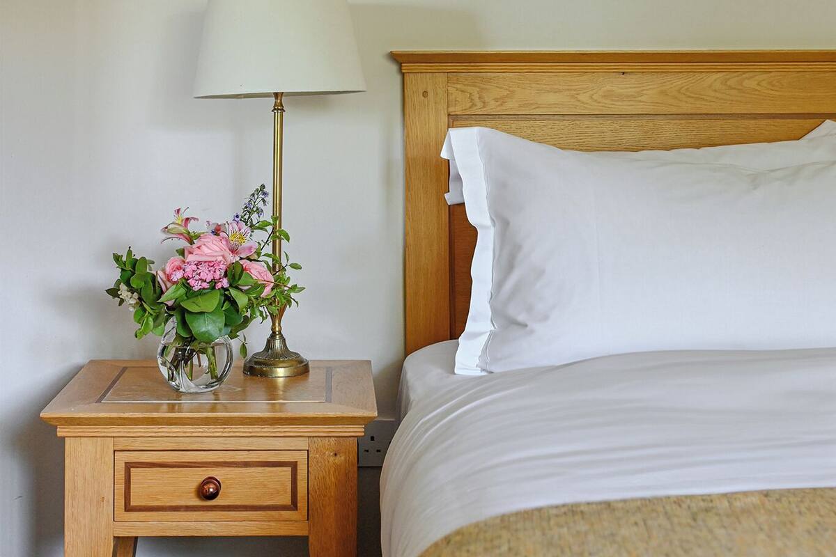 Una cama bien hecha con sábanas blancas, una mesita de noche de madera con una lámpara y un jarrón de flores, y un cuadro en la pared.