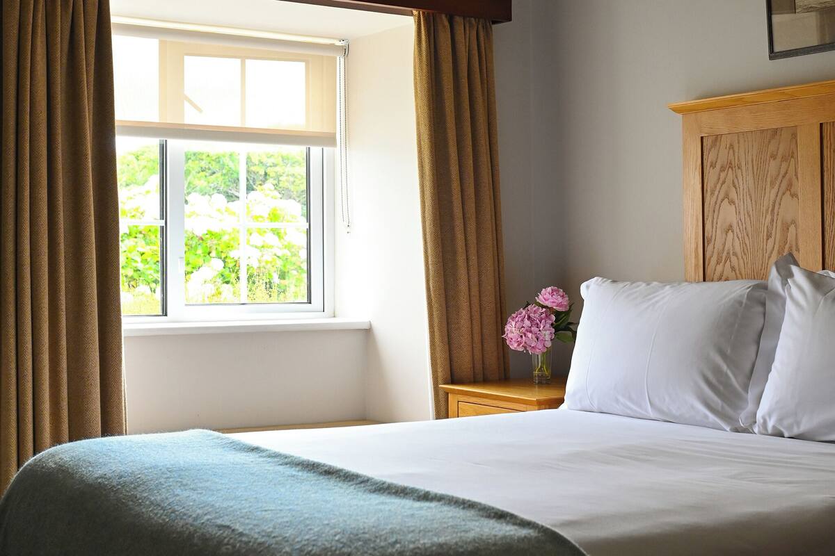 Una cama bien hecha con sábanas blancas, cabecera de madera y una ventana con cortinas.