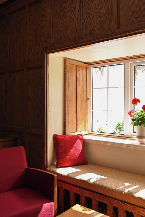 Una habitación con un revestimiento de madera, una ventana con vistas a follaje, un banco con cojín rojo y una silla roja.