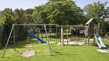 Kids can play on swings, slides, and a climbing frame in this outdoor playground surrounded by a lush lawn and trees.
