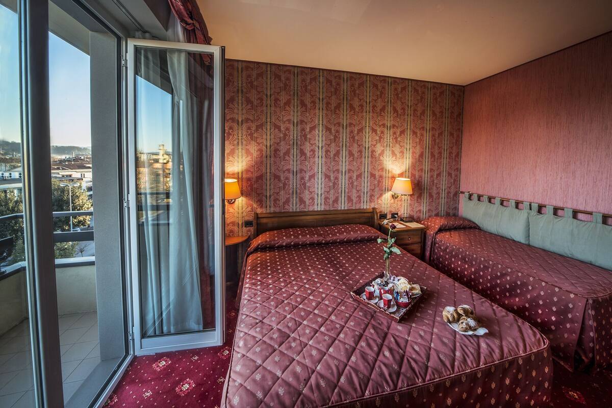 A hotel room with a large window, a bed with a quilted cover, two bedside tables with lamps, and a view of a cityscape.