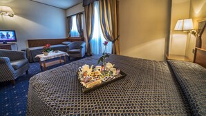 Una habitación de hotel con una cama grande, dos sillones, una mesita con una bandeja de desayuno, un televisor y cortinas.