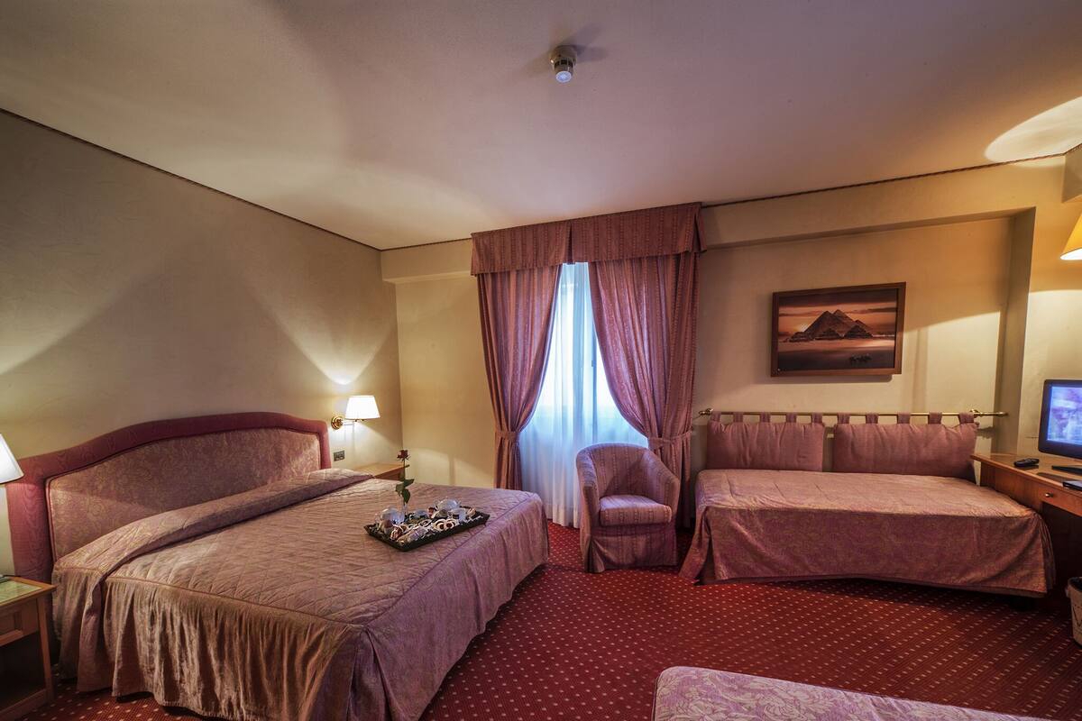 A hotel room with a large bed, a smaller bed, a chair, a desk with a computer, and a window with curtains.