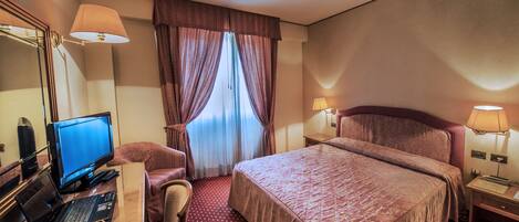 Une chambre d’hôtel avec un lit, un bureau avec un ordinateur portable, une chaise, une lampe de bureau et une fenêtre avec des rideaux.