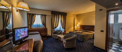 A hotel room with a bed, two armchairs, a desk with a laptop, a television, and a window with curtains.