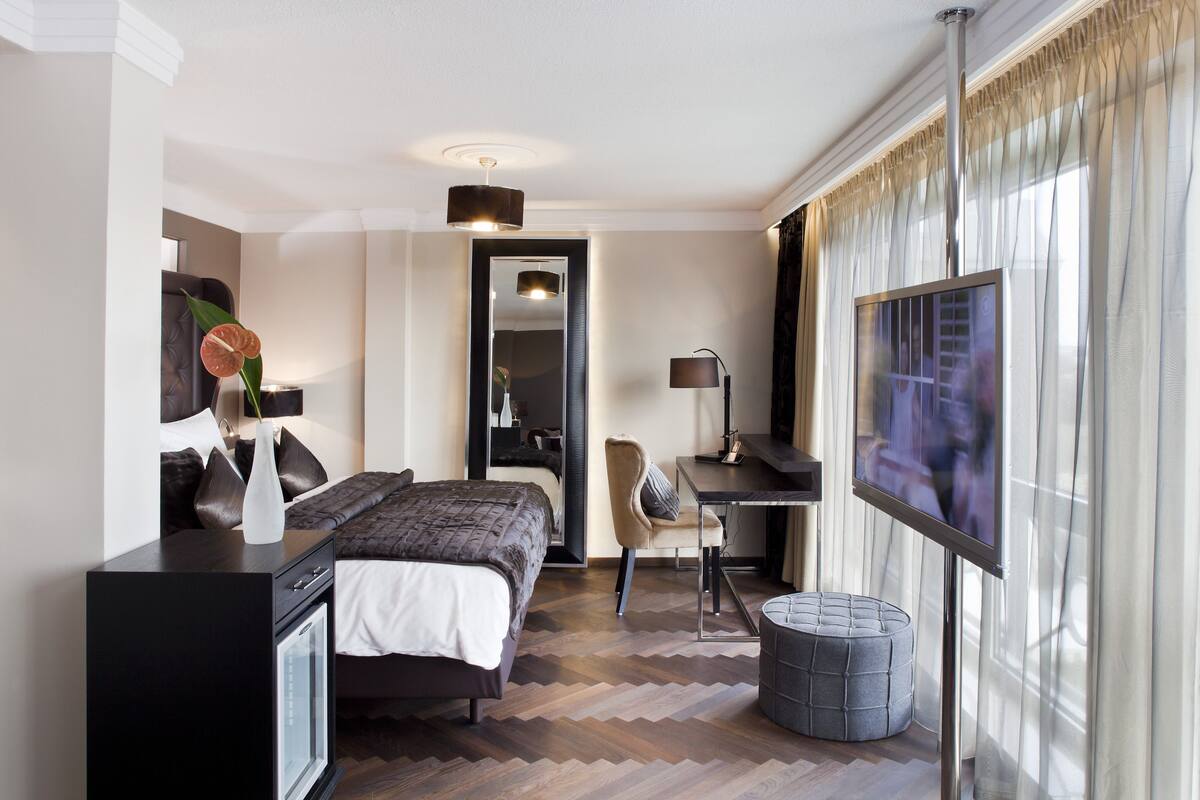 A modern hotel room with a bed, desk, chair, TV, and a small ottoman.
