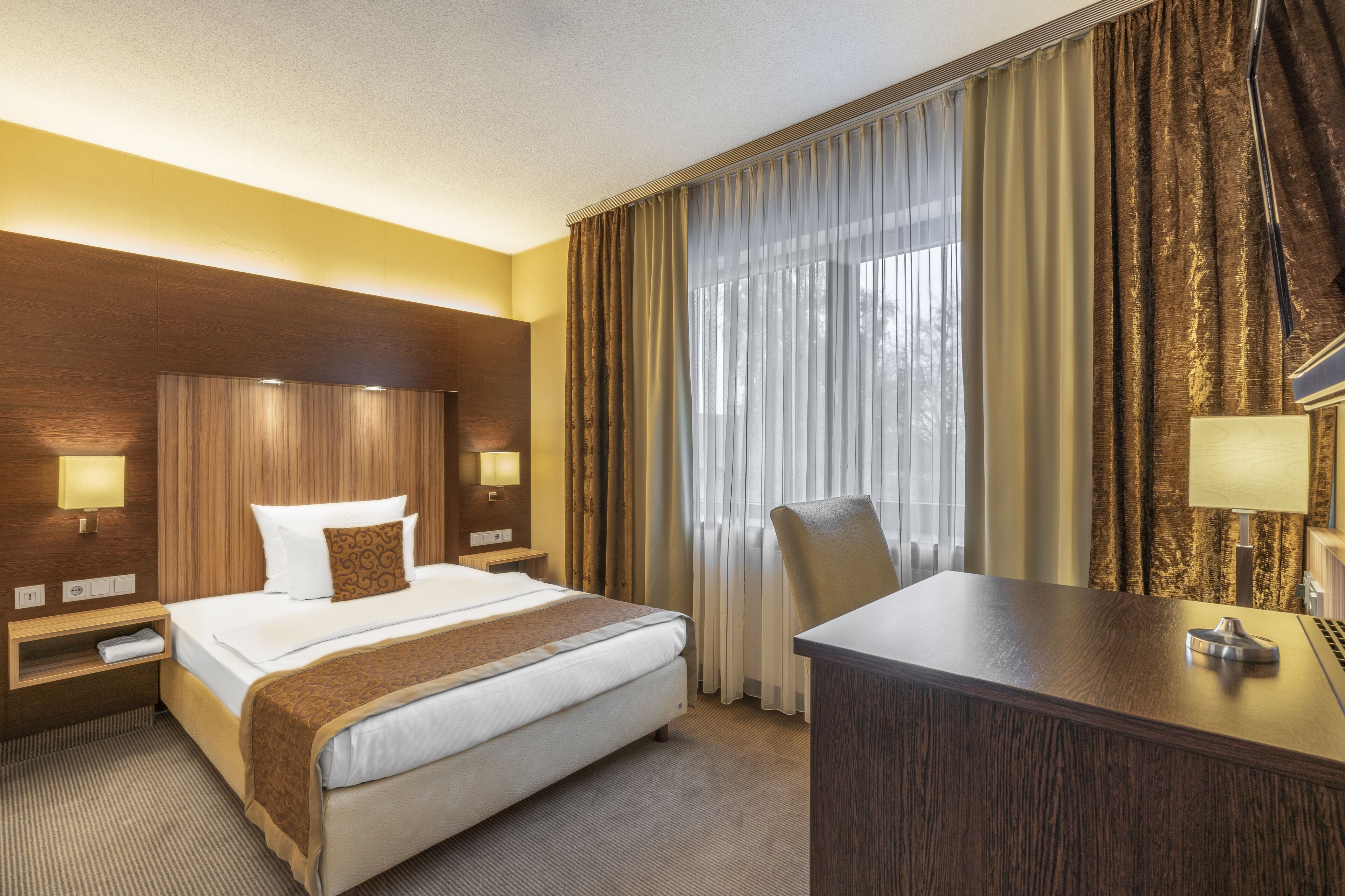 A hotel room with a large bed, a desk, and a chair. There are two bedside tables with lamps, a window with curtains, and a wooden headboard.