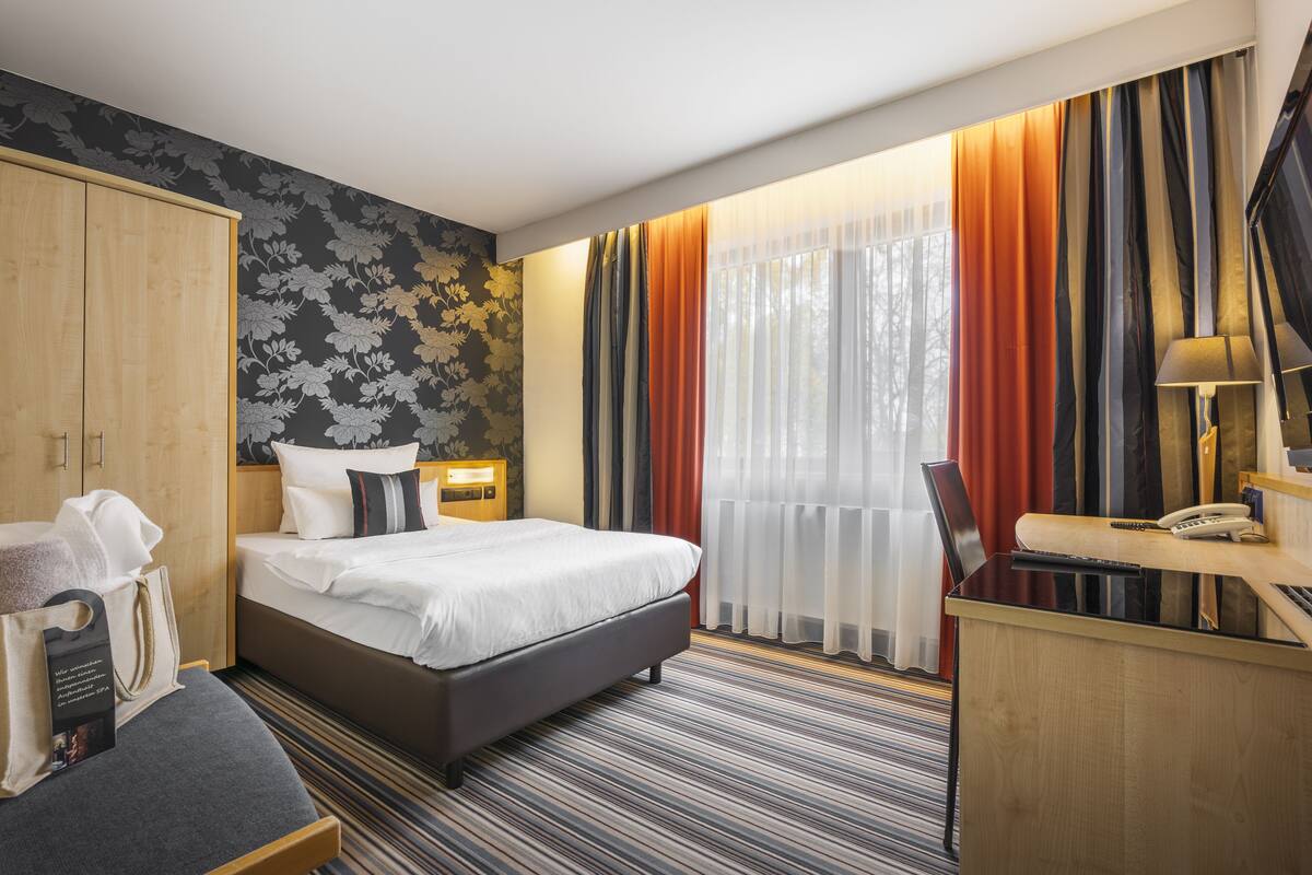 A hotel room with a bed, a desk, a wardrobe, and a window with curtains.
