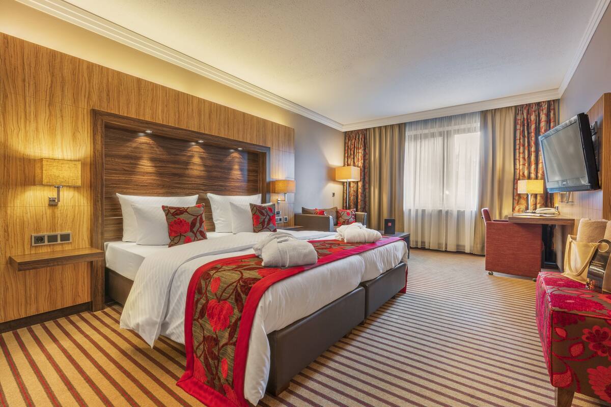 A hotel room with a large bed, a desk, a chair, a TV, and a window with curtains.