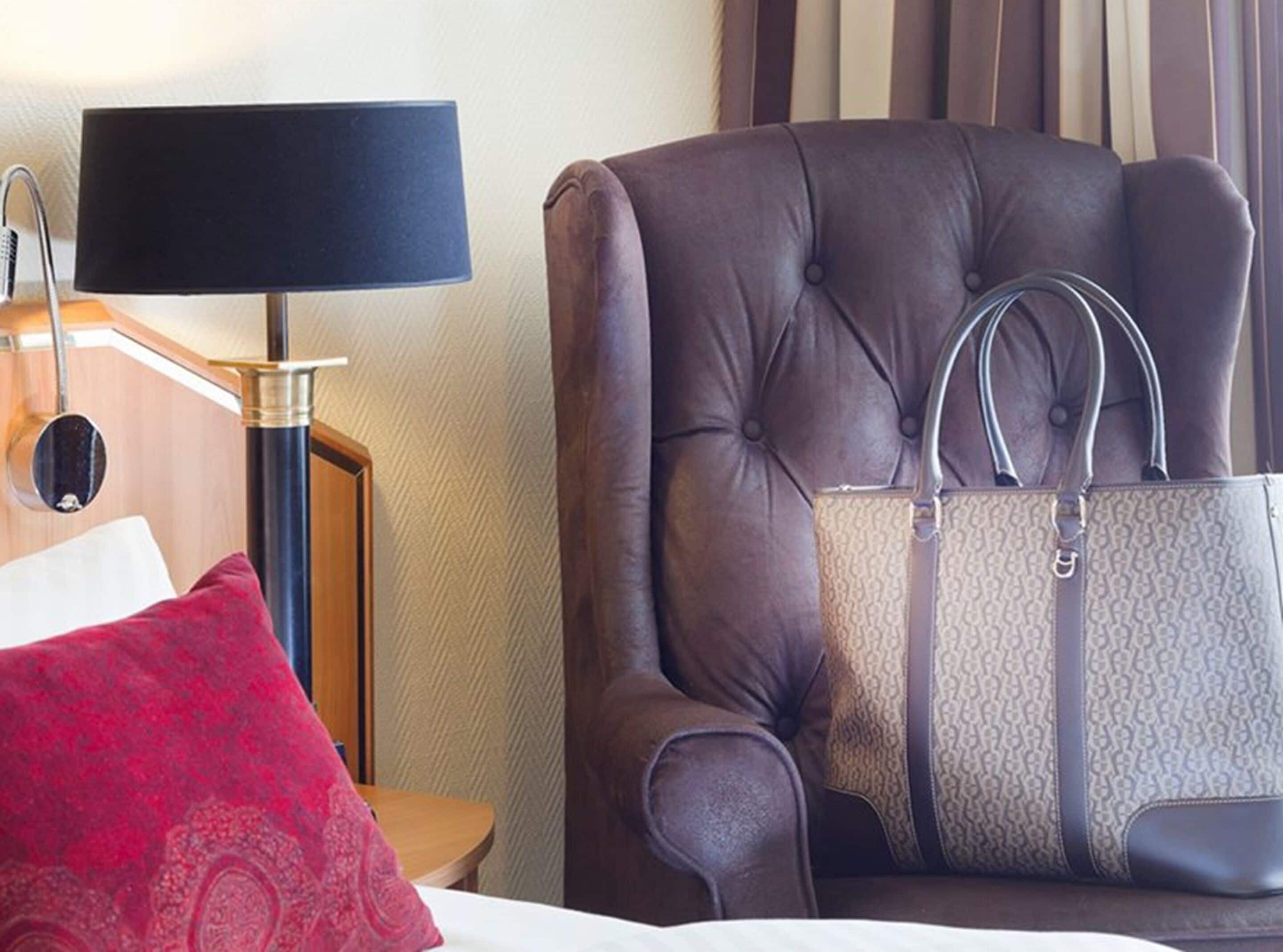 A hotel room with a large brown leather armchair, a matching handbag, a red pillow, and a lamp on a wooden side table.