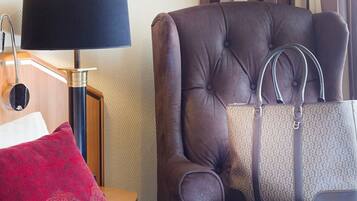 Habitación de hotel con un sillón grande de cuero marrón, un bolso a juego, un cojín rojo y una lámpara sobre una mesita de noche de madera.