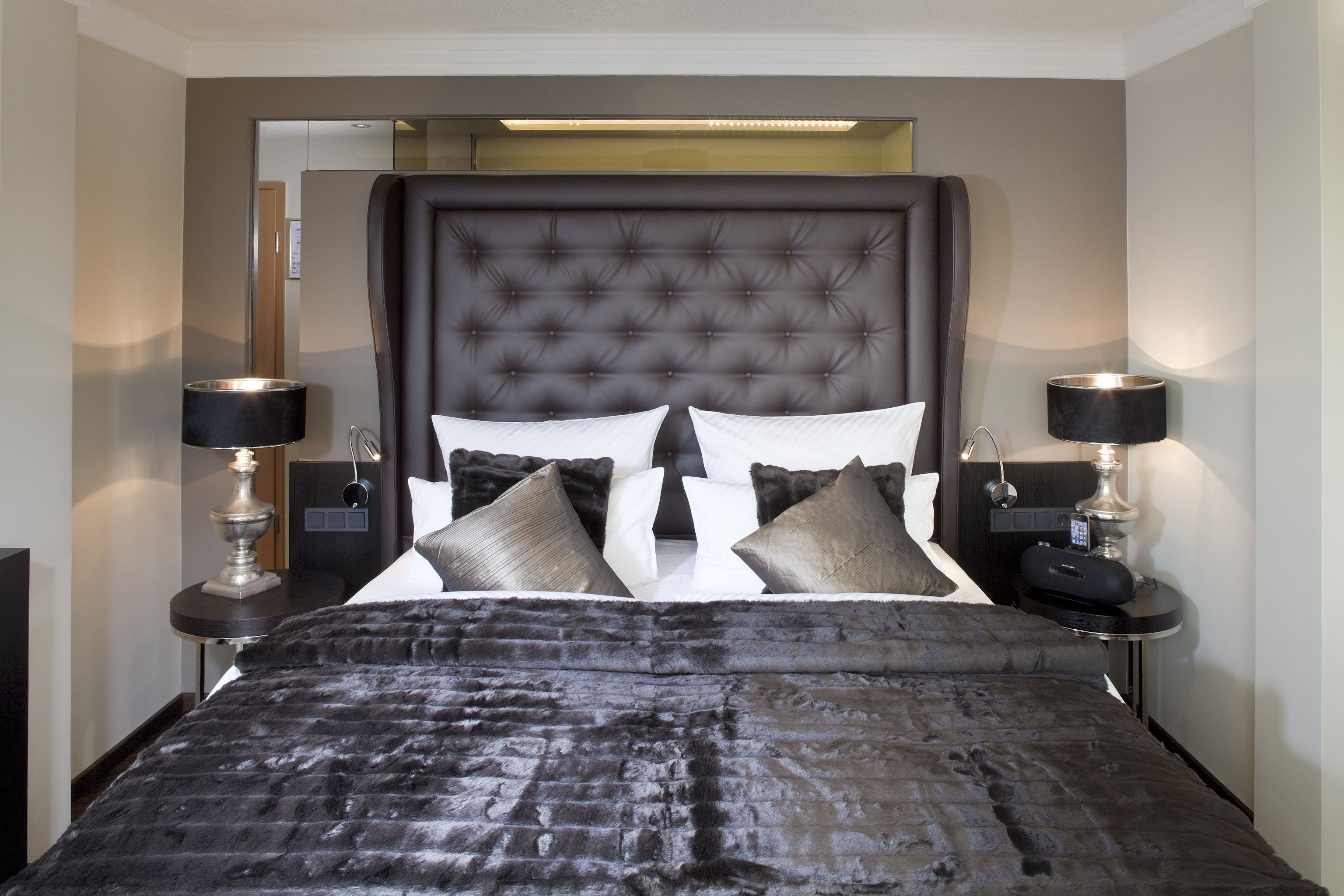 Une chambre d’hôtel moderne avec une tête de lit capitonnée, un grand lit recouvert d’une couverture à motifs foncés, deux tables de chevet avec des lampes et un miroir posé sur l’une des tables.