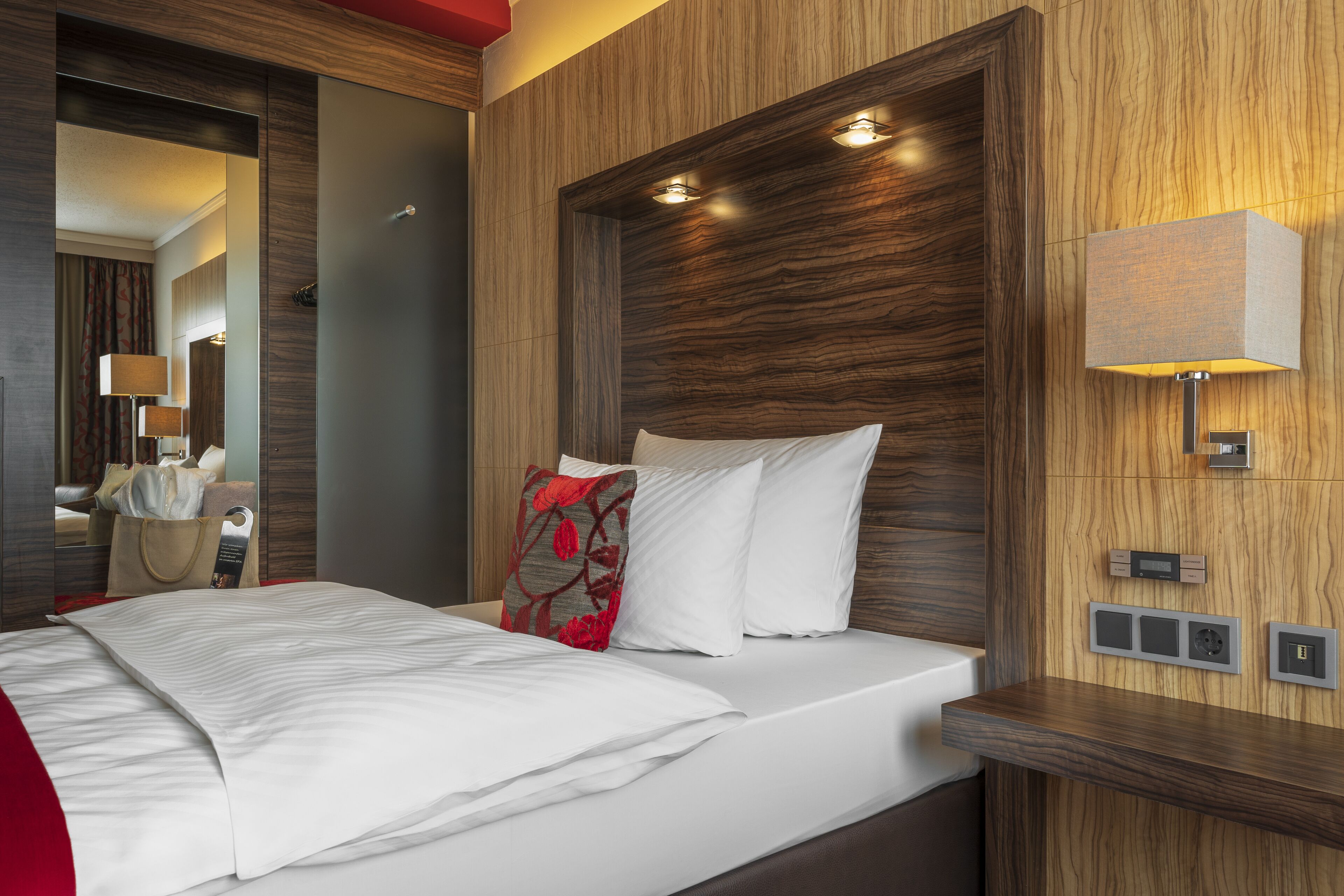 A hotel room with a large bed, a bedside table, a mirror, and a wall-mounted lamp.