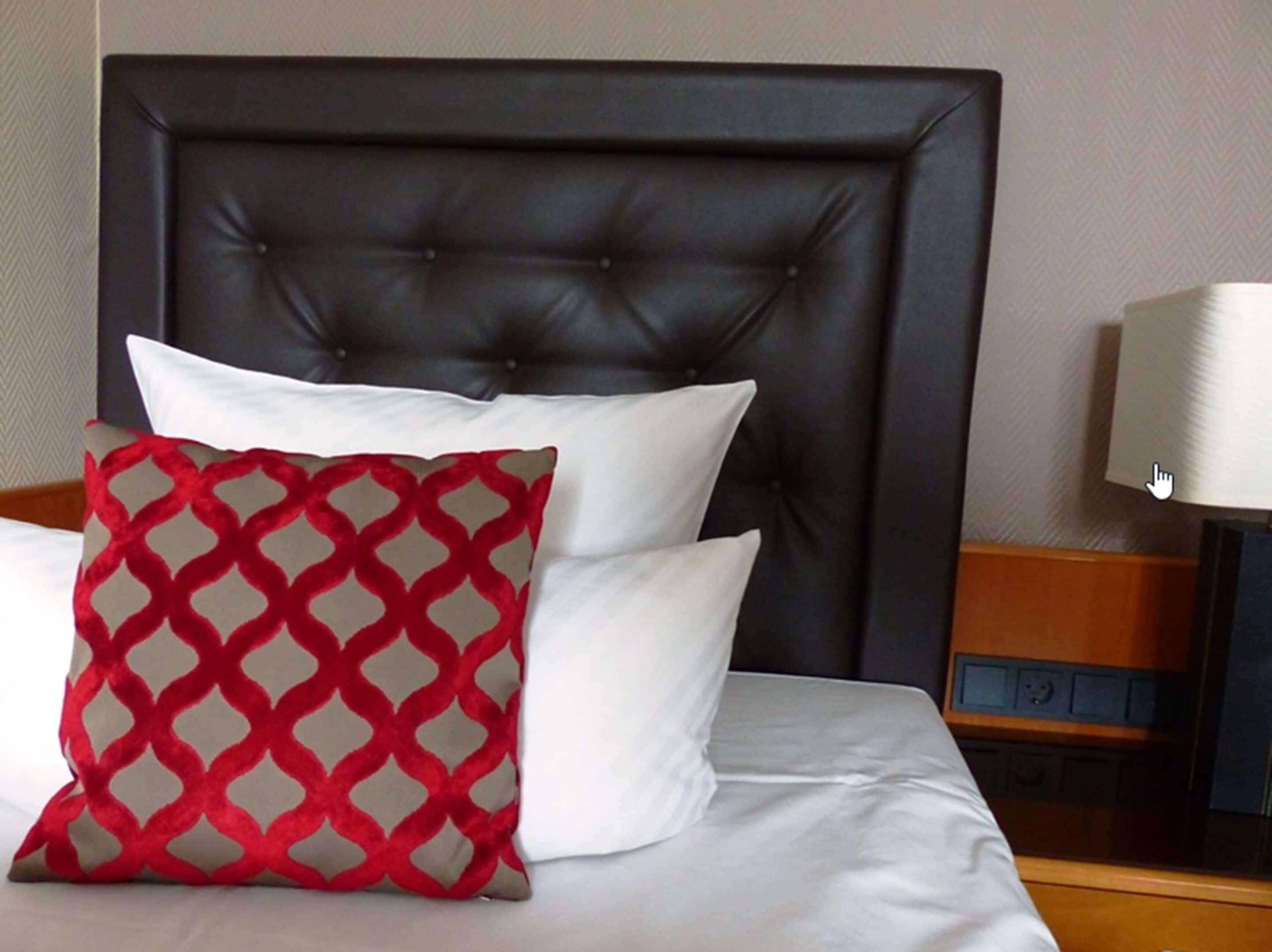 Un lit avec une tête de lit noire recouverte de tissu, des oreillers blancs et un coussin à motifs rouges et beiges.
