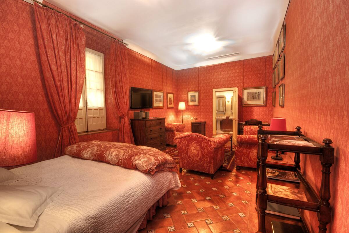 Una habitación con cama, una silla, un banco de madera, cómoda, televisión y un espejo en la pared.