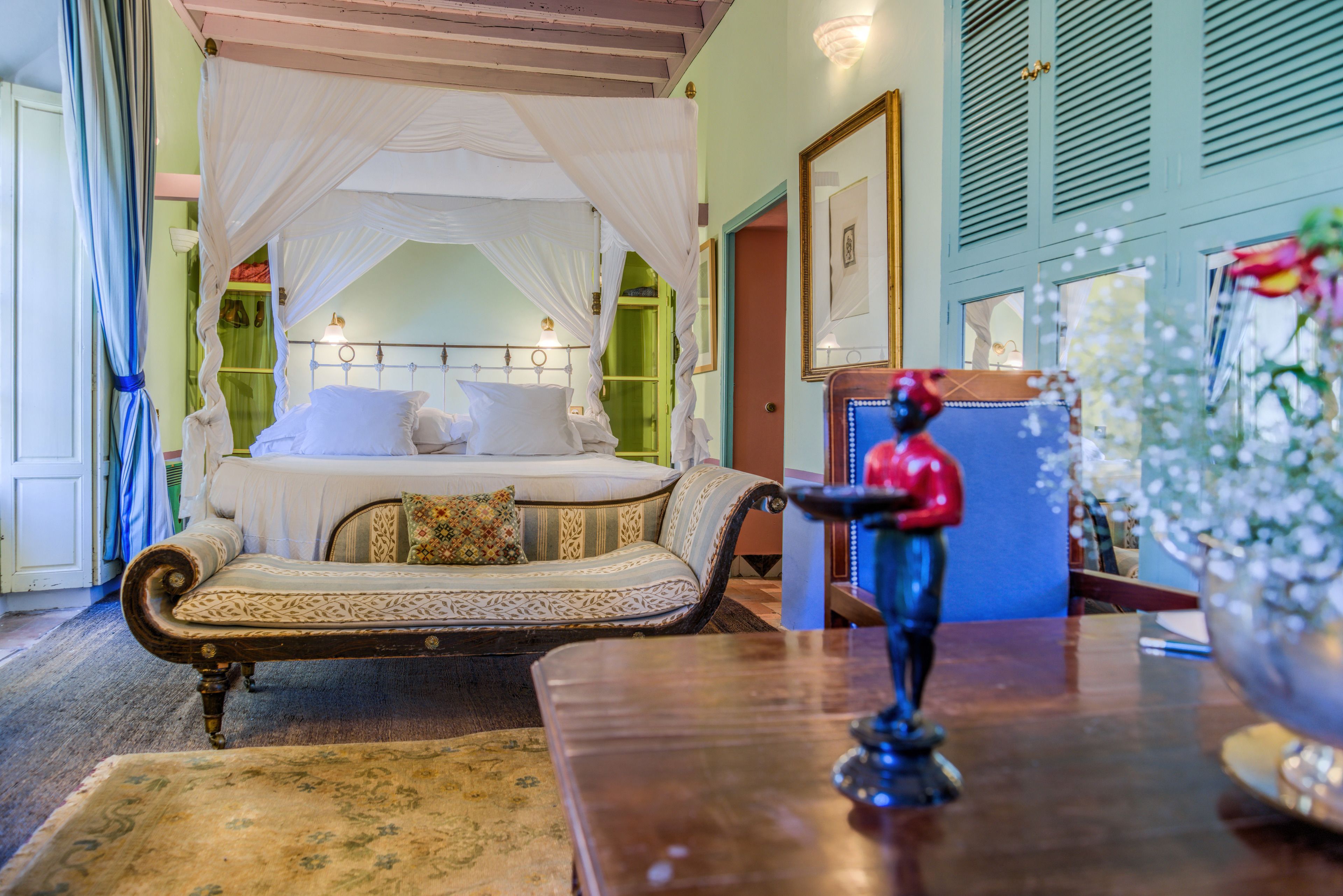 Un dormitorio con cama con dosel, un sofá, una mesita con una estatua y un jarrón con flores.