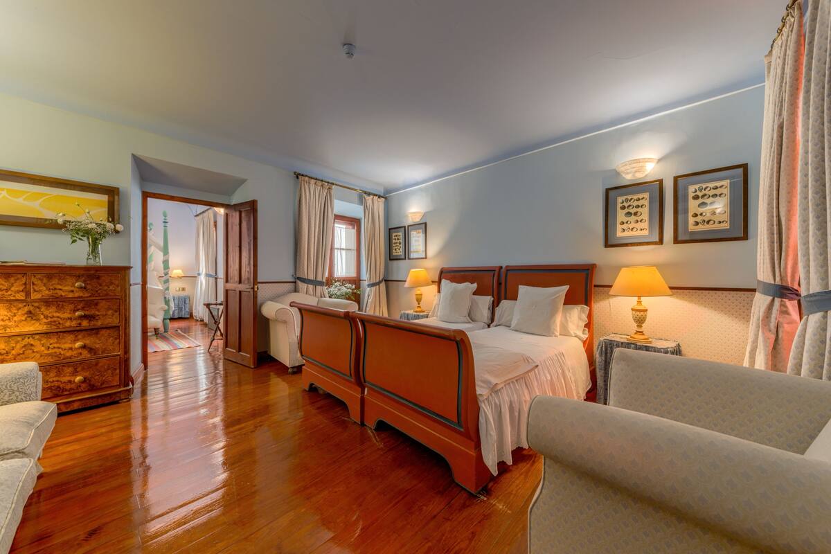 Habitación de hotel con piso de madera, cama con ropa blanca, dos mesitas de noche con lámparas, cómoda y una silla.