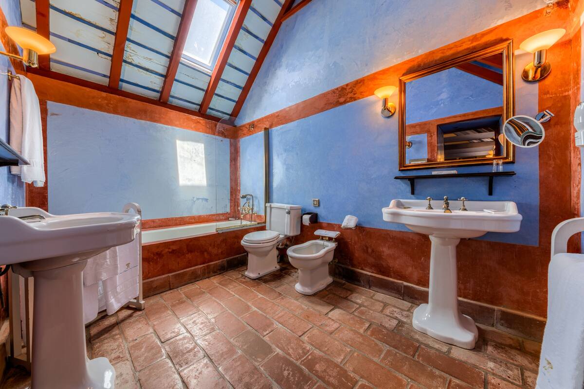Un baño con tragaluz, un espejo grande, dos inodoros y un lavamanos.