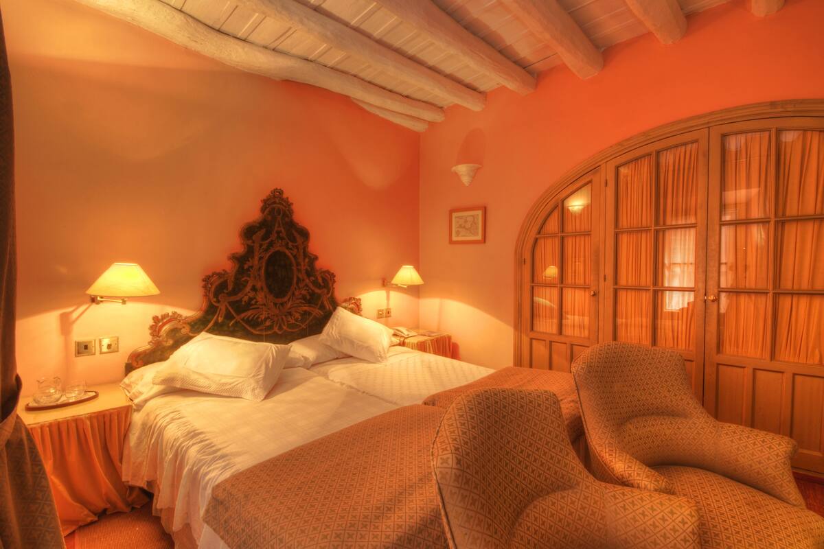 Una habitación de hotel con una cama grande, dos lámparas de noche, un armario de madera y un sillón estampado.