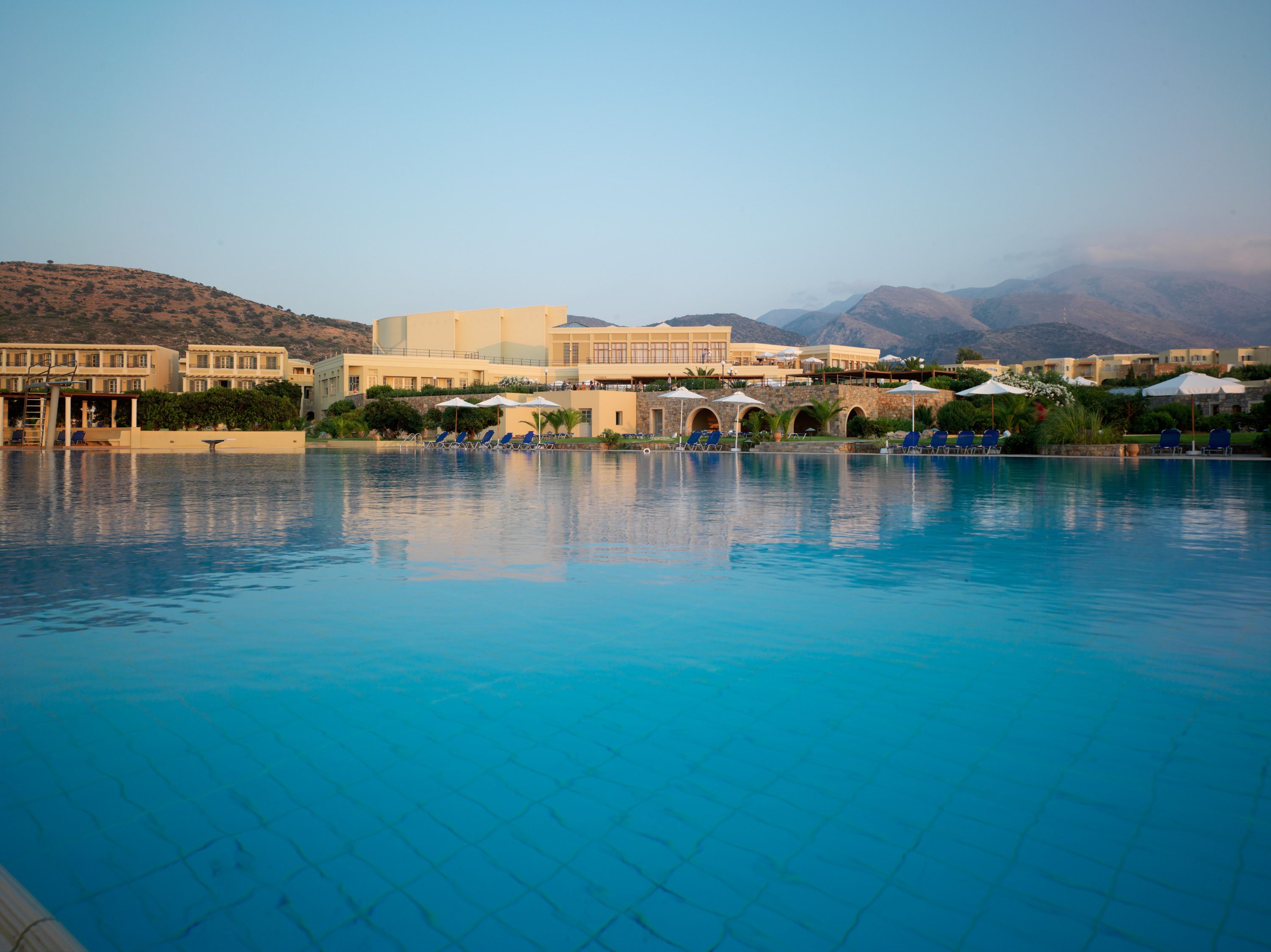 A swimming pool with lounge chairs and umbrellas, surrounded by buildings and mountains.