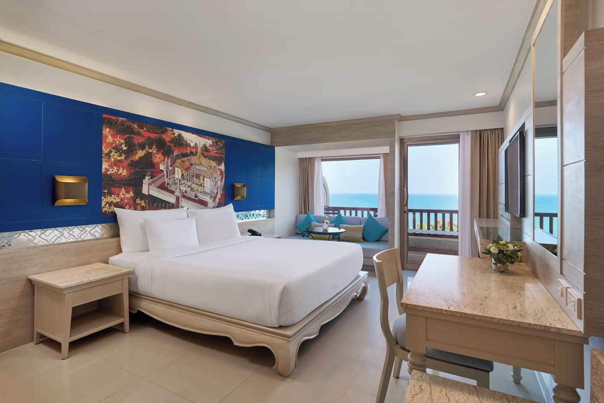 Une chambre d’hôtel avec un lit, une table de chevet, un bureau, une chaise et un grand tableau au mur.