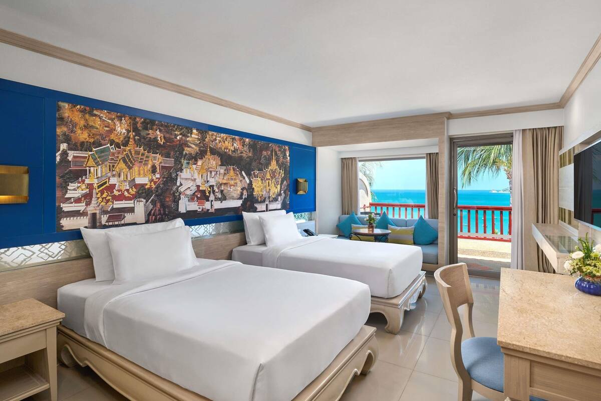 A hotel room with two beds, a desk, a chair, a TV, and a balcony with a view of the sea.
