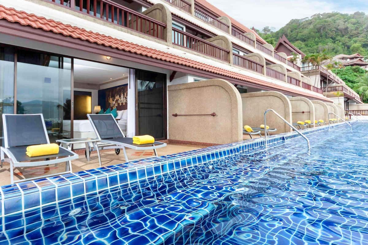 A hotel with a swimming pool, sun loungers, and a view of a forested hill.