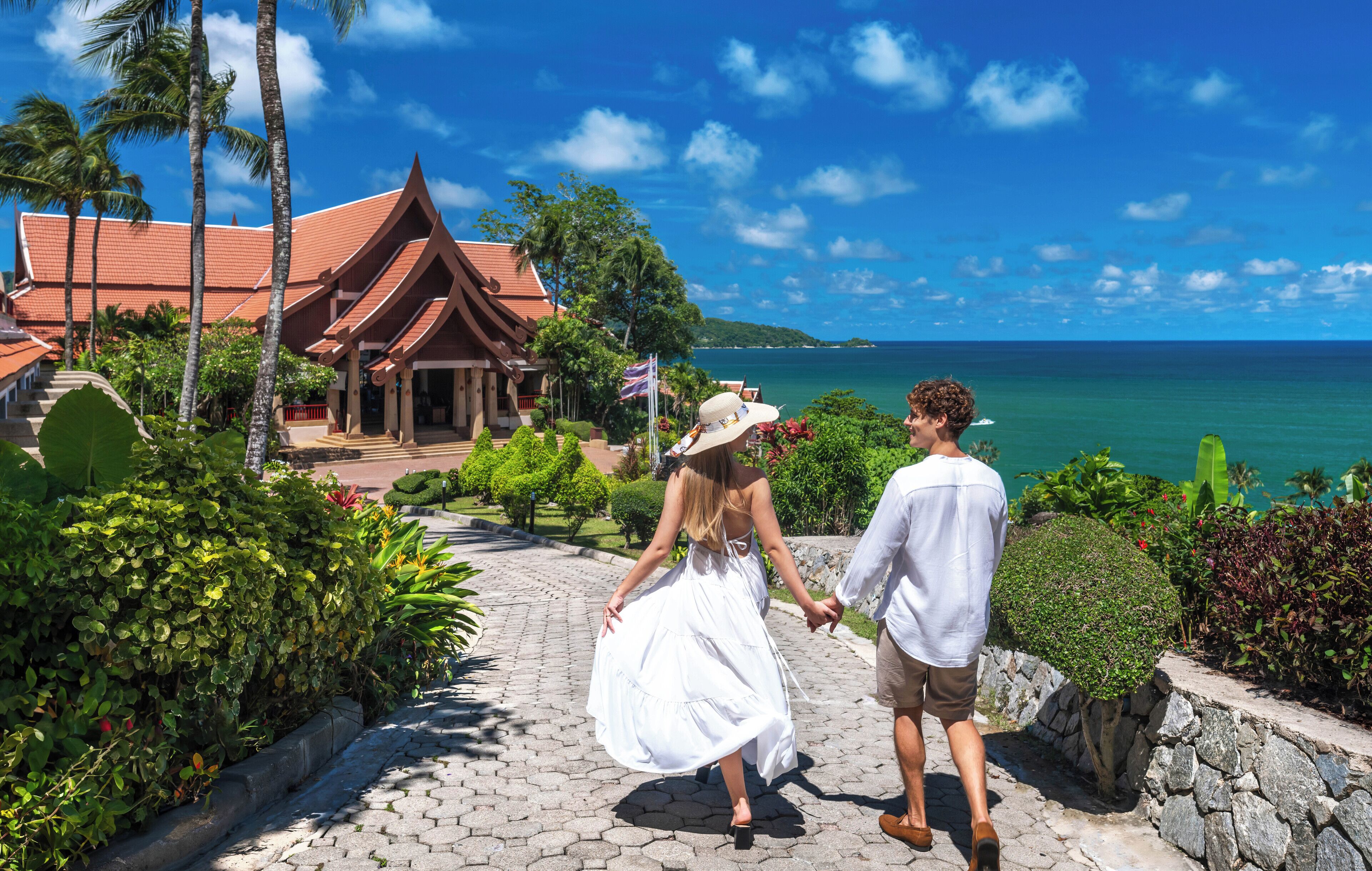 A couple walking on a paved pathway with a view of a traditional building and the ocean.