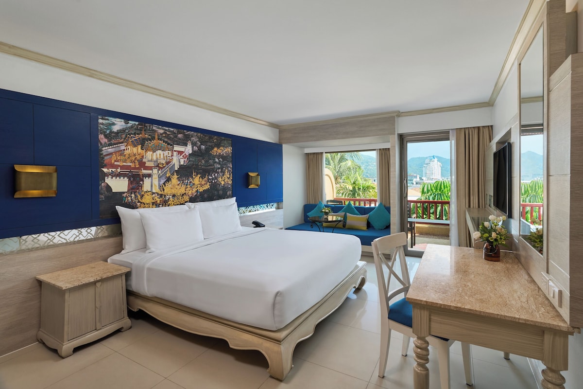 Une chambre d’hôtel avec un grand lit, un bureau, une chaise, une télévision et un balcon offrant une vue sur la verdure et les bâtiments.