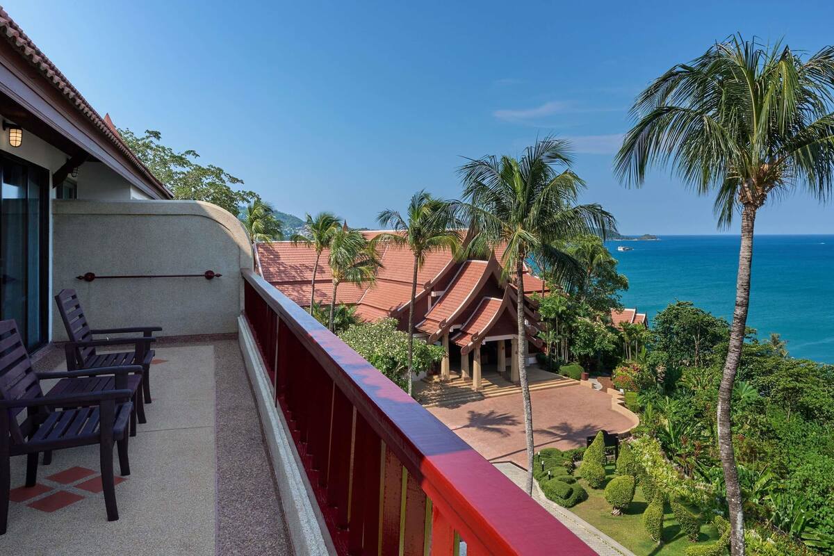 Ein Balkon mit Stühlen, Blick auf den Ozean, Palmen und ein Gebäude mit rotem Dach.