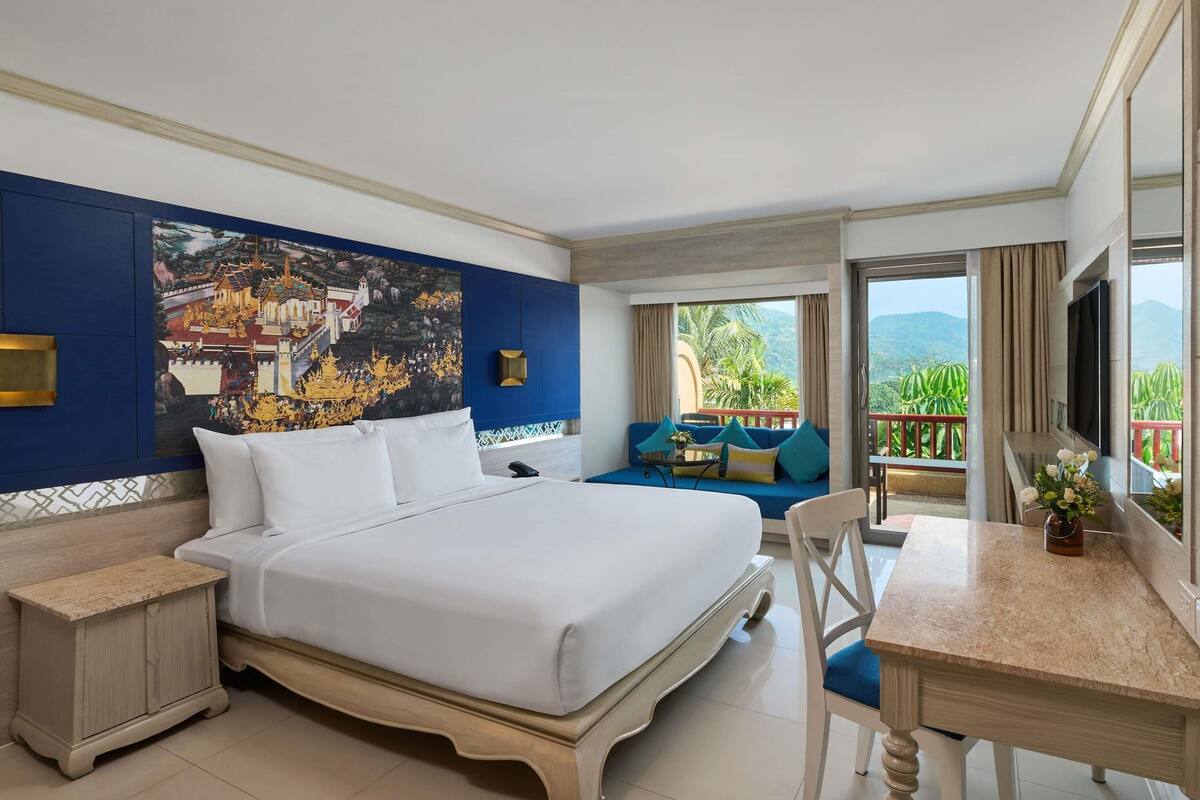 A hotel room with a large bed, a sofa, a dining table, and a balcony with a view.