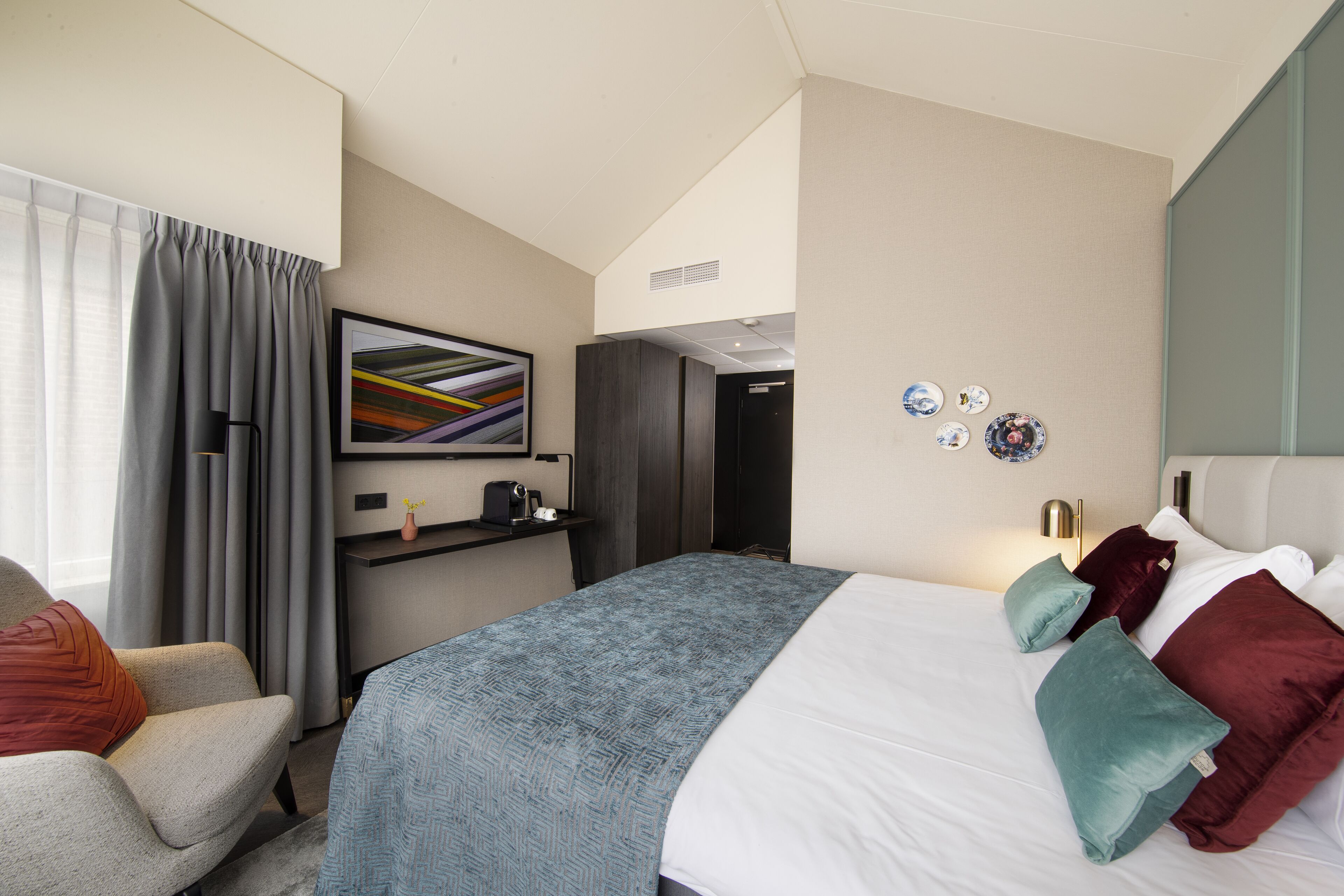 A modern hotel room with a large bed, a desk with a chair, a window with curtains, and a wall-mounted artwork.
