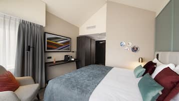 A modern hotel room with a large bed, a desk with a chair, a window with curtains, and a wall-mounted artwork.