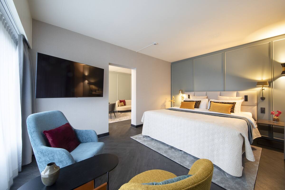 A modern hotel room with a large bed, a seating area with a chair and table, a flat-screen TV mounted on the wall, and a large window with curtains.