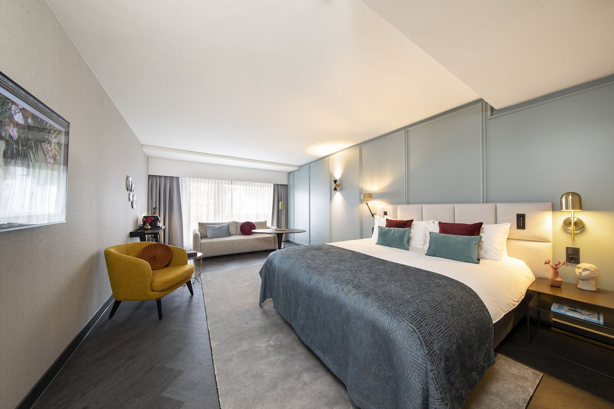 A modern hotel room with a large bed, a sofa, a chair, and a desk.