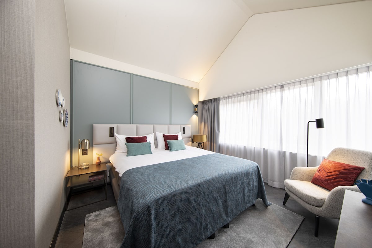 A modern hotel room with a large bed, a nightstand with a lamp, a chair, and a view of the outdoors through sheer curtains.