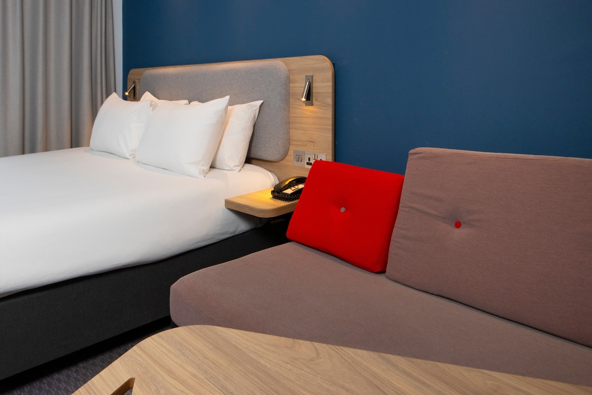 A hotel room with a bed, a sofa, a wooden nightstand, and a red laptop.