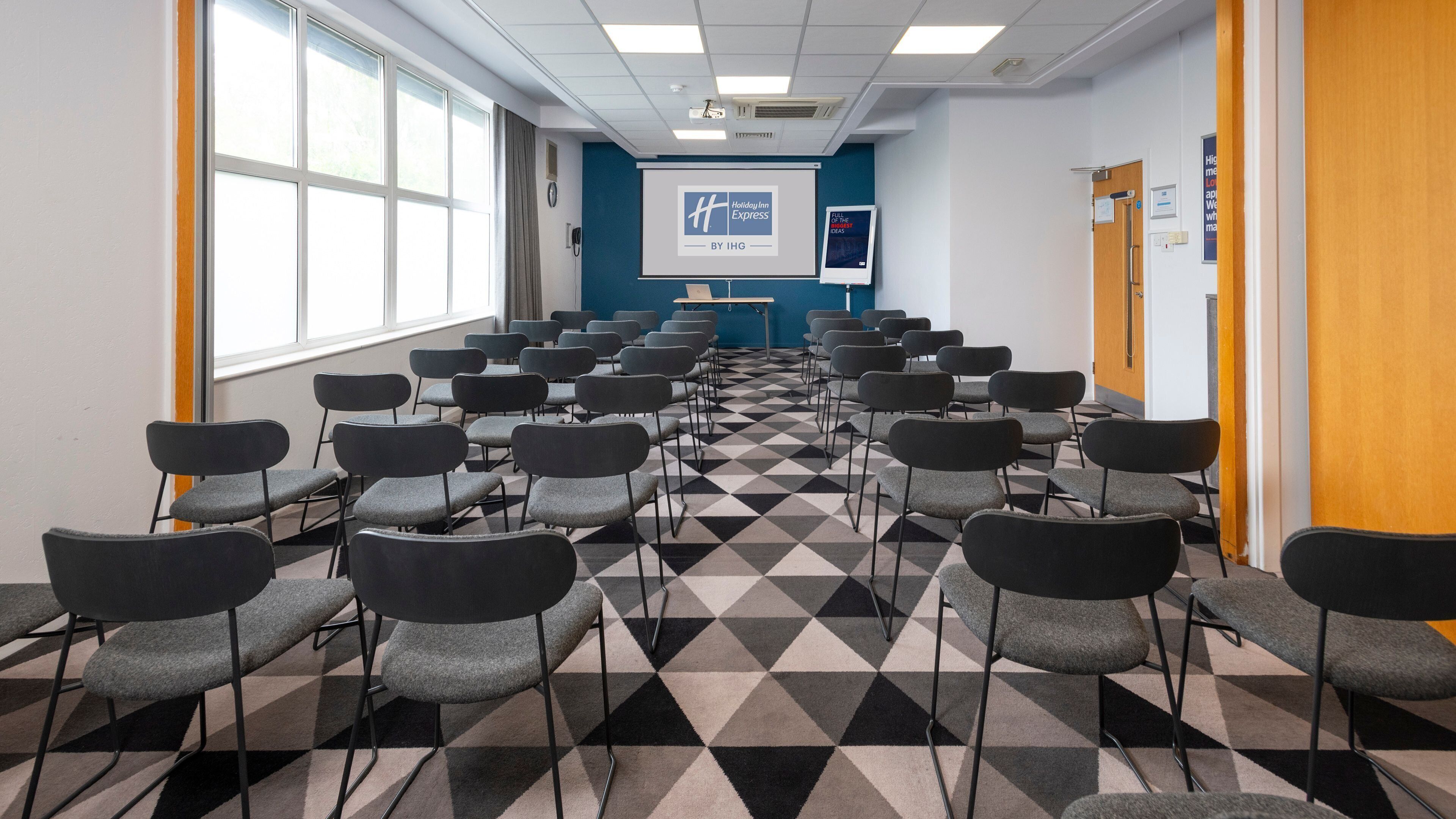 A conference room with rows of chairs, a projector screen, and a presentation on the wall.