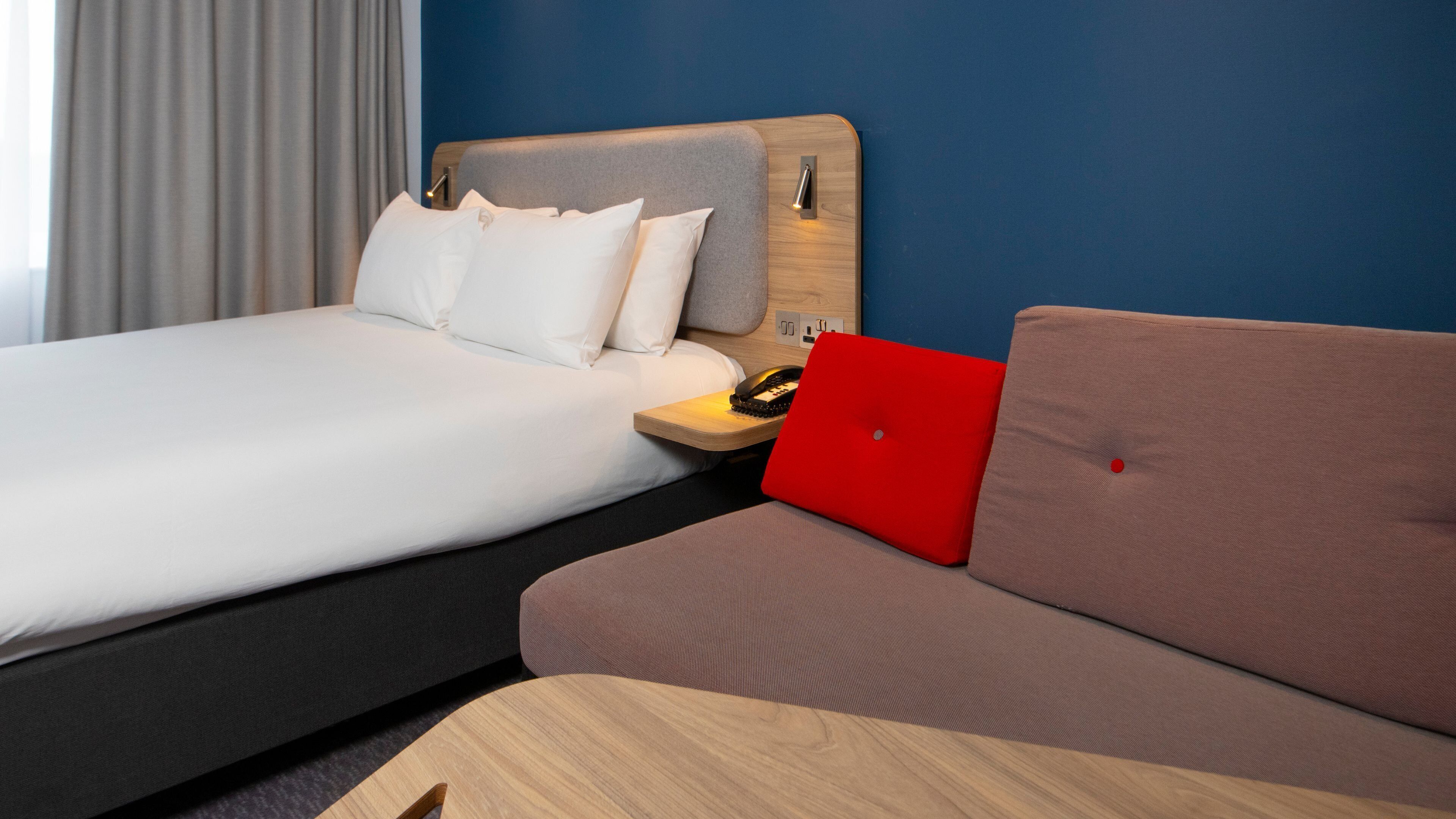 A hotel room with a bed, a sofa, a wooden nightstand, and a red laptop.