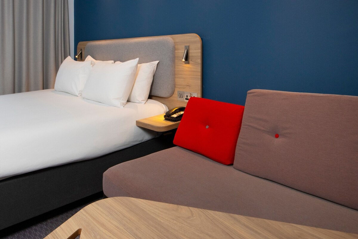 Una habitación de hotel con una cama, un sofá, una mesita de noche de madera y un ordenador portátil rojo.