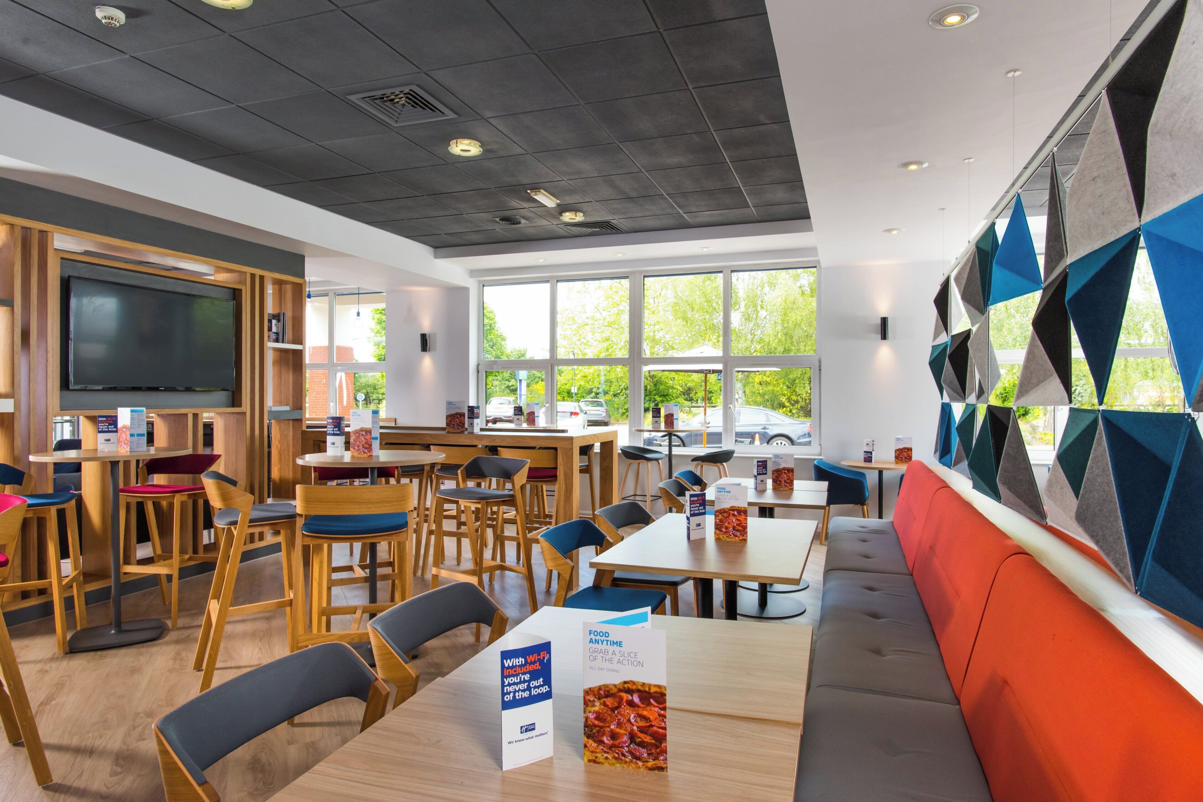 A modern restaurant with wooden furniture, a flat-screen TV, and large windows offering a view of the outdoors.