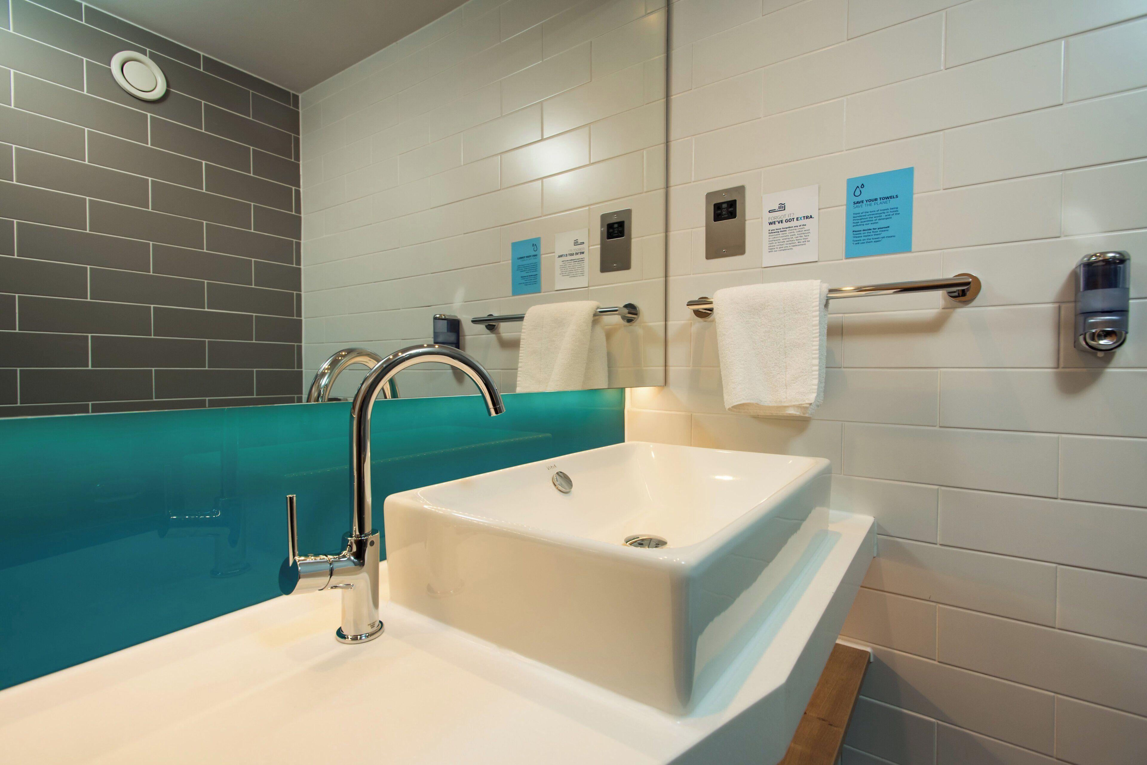 A modern bathroom with a white sink, a chrome faucet, and a teal-colored bathtub. There are towels hanging on a rack and a sign on the wall.