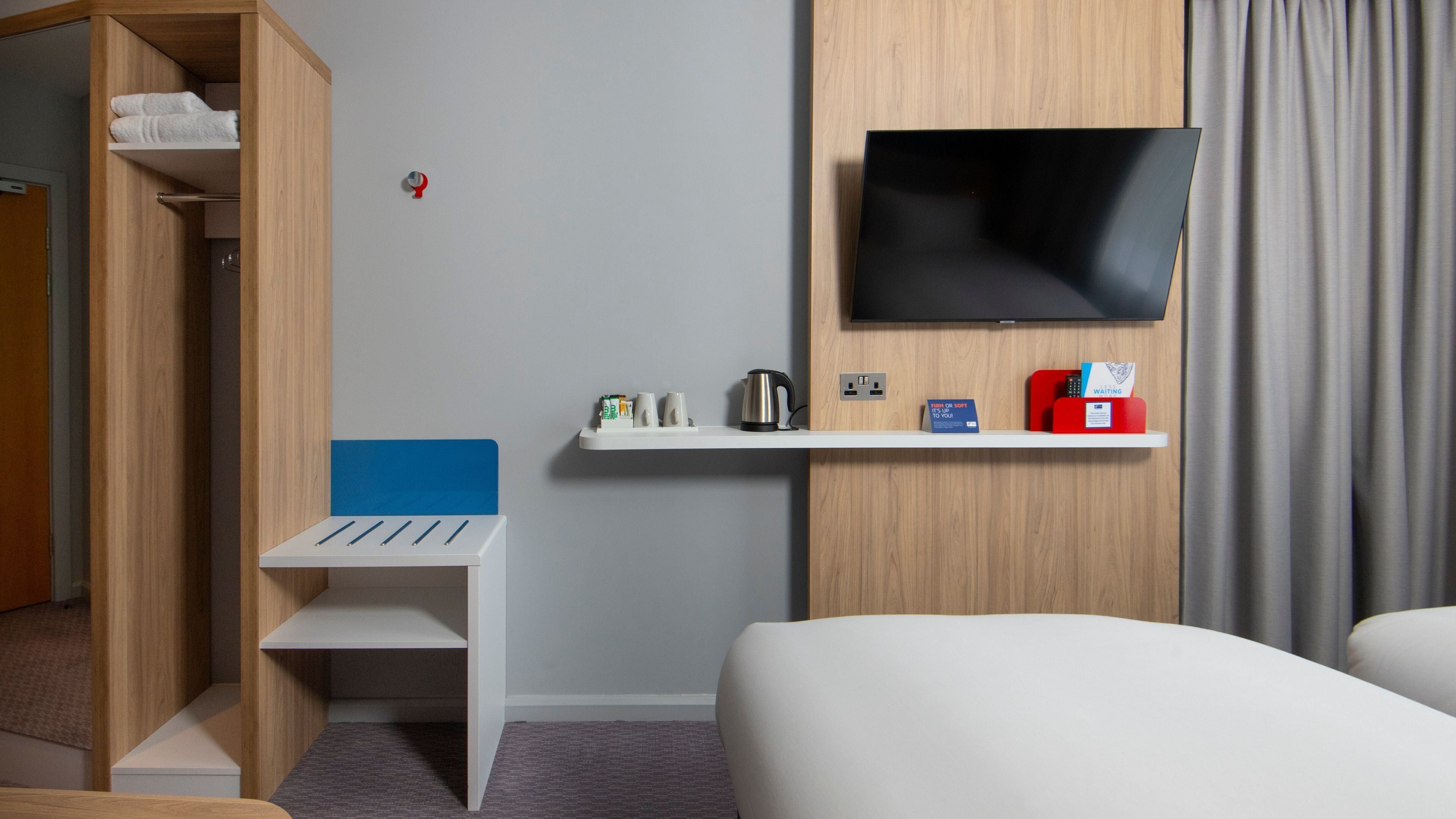 A hotel room with a bed, a TV mounted on the wall, a small table with a kettle, and a shelf with a red box and a sign.