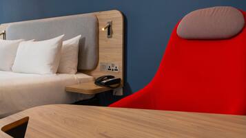 A hotel room with a bed, a red chair, a bedside table with a phone, and a wall socket.
