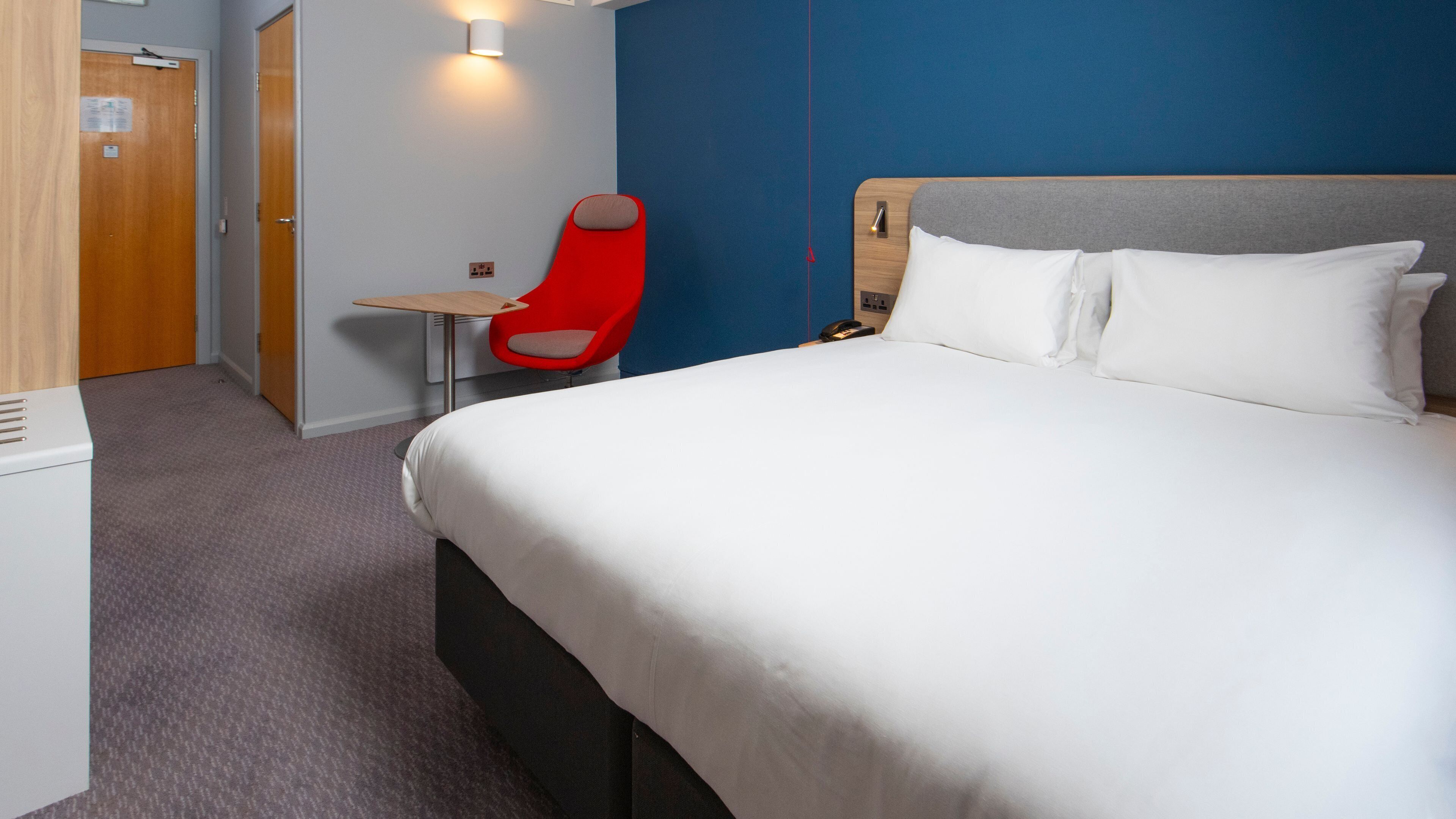 A hotel room with a bed, a desk, a chair, and a wall-mounted light.