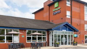 A Holiday Inn Express building with a blue entrance, outdoor seating area, and a sign welcoming guests. - Holiday Inn Express Burton Upon Trent by IHG (Burton on Trent)