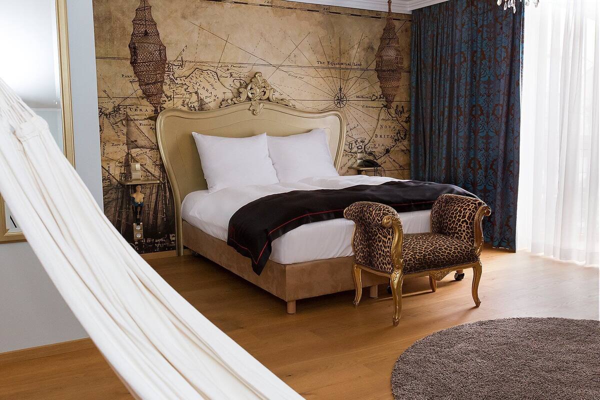 Une chambre à coucher avec un grand lit, une chaise longue, une carte accrochée au mur et une fenêtre ornée de rideaux.