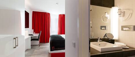 A modern hotel room with a bed, a desk, and a bathroom with a sink and mirror.